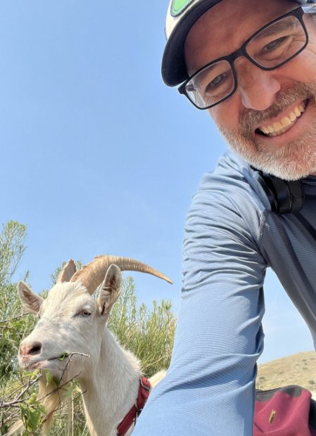 A smiling hiker beside Krampus, a horned goat browsing on a sunny Colorado trail
